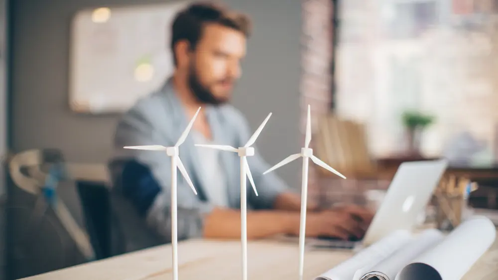 Ein Mann sitzt an seinem Schreibtisch und plant ein Windkraftprojekt, während im Vordergrund auf dem Tisch drei Windkrafträdermodelle stehen.