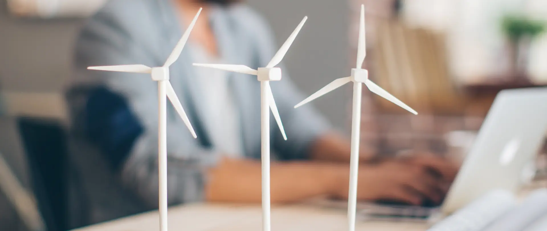 Ein Mann sitzt an seinem Schreibtisch und plant ein Windkraftprojekt, während im Vordergrund auf dem Tisch drei Windkrafträdermodelle stehen.