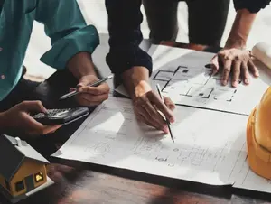 Bauplanung Bauplaner sitzen und stehen am Tisch. Einer von ihnen hält einen Taschenrechnern der andere zeichnet etwas in den Plan ein