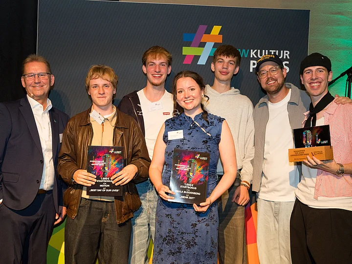 WWKulturSTARTER26 - Finalist*innen Gruppenfoto aller ausgezeichneten KulturSTARTER mit Urkunden und Auszeichnungen vor der Fotowand der Preisverleihung.