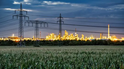 Netzersatzanlage für Industrie und Gewerbe zur Notstromversorgung Strommasten und Hochspannungsleitungen führen zu einer beleuchteten Industrieanlage bei Dämmerung.