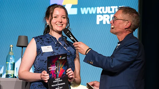 Paula Buddenberg - Start-Bonus Paula Buddenberg hält eine Urkunde für den Start-Bonus des WWKulturSTARTER26 in der Hand, während der Moderator Ingo Börchers ein Interview auf der Bühne führt.