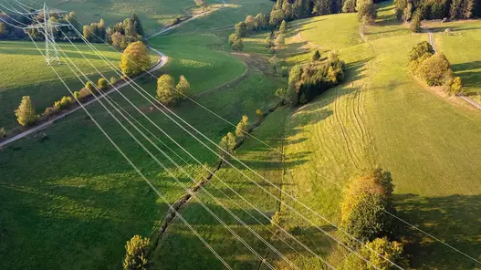 Verteilnetze für eine sichere Energieversorgung Das Bild zeigt Stromleitungen, die sich durch eine grüne, ländliche Landschaft mit Wiesen und Hügeln ziehen.