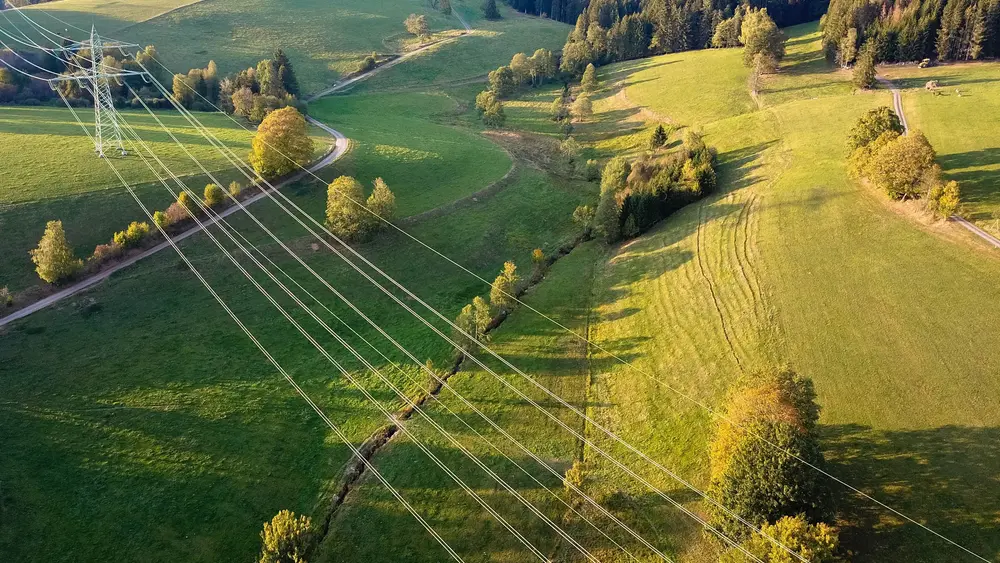 Das Bild zeigt Stromleitungen, die sich durch eine grüne, ländliche Landschaft mit Wiesen und Hügeln ziehen.