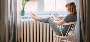 Frau mit Notizbuch am Fenster Eine Frau sitzt auf einem Holzstuhl am Fenster und hat die Füße auf der Heizung. Sie hält ein Bleistift sowie ein Notizbuch in den Händen