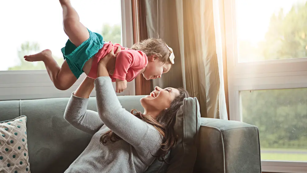 Eine Mutter liegt auf dem Sofa in einem gemütlichem Wohnzimmer. Die Mutter spielt mit der Tochter und beide sind sehr glücklich, Die Sonne scheint durch die großen Fenster rein,