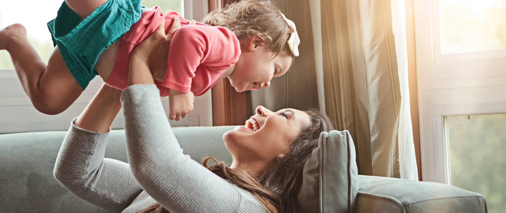 Eine Mutter liegt auf dem Sofa in einem gemütlichem Wohnzimmer. Die Mutter spielt mit der Tochter und beide sind sehr glücklich, Die Sonne scheint durch die großen Fenster rein,