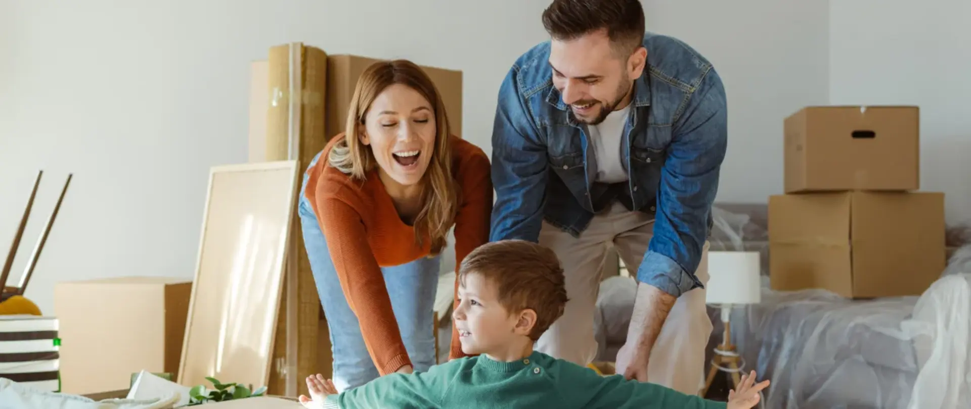 Eine Familie mit kleinem Sohn schiebt zwischen Kartons ihren Sohn im Karton durch das neue Wohnzimmer