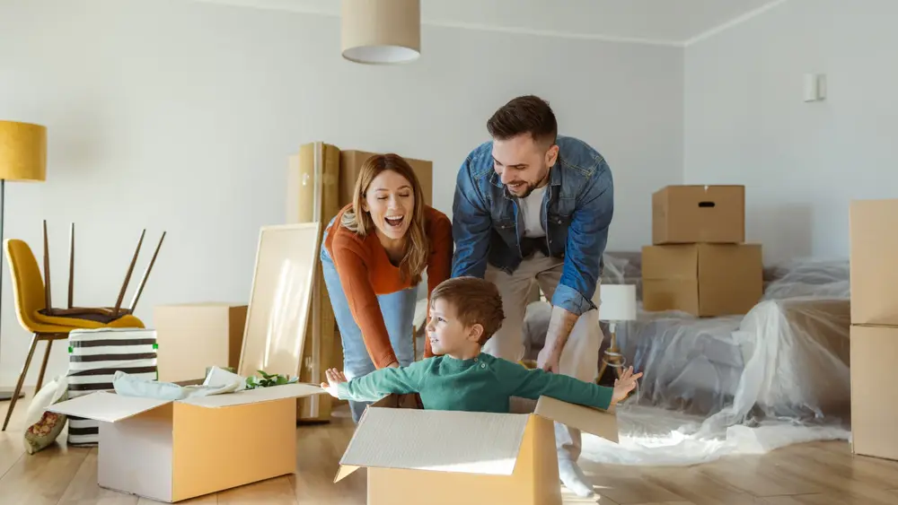 Eine Familie mit kleinem Sohn schiebt zwischen Kartons ihren Sohn im Karton durch das neue Wohnzimmer