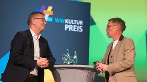 WWKulturpreis Unterhaltung Zwei Männer stehen an einem Stehtisch vor einem großen Banner und unterhalten sich