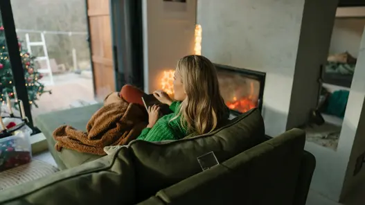Gemütliche Stimmung zu Hause Eine Frau sitzt im gemütlichen Wohnzimmer auf ihrem Sofa und schaut nach draußen. Der Kamin ist an und es ist ein kleiner Teil eines Weihnachtsbaums zu sehen