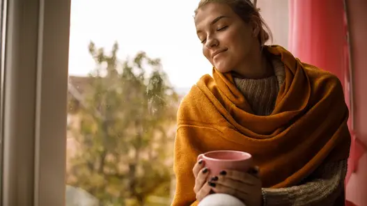 Wärme genießen Eine in der Decke eingewickelte Frau sitzt auf einer Fensterbank, hält eine Tasse Kaffee in den Händen und genießt den Moment. Ihre Augen sind geschlossen.