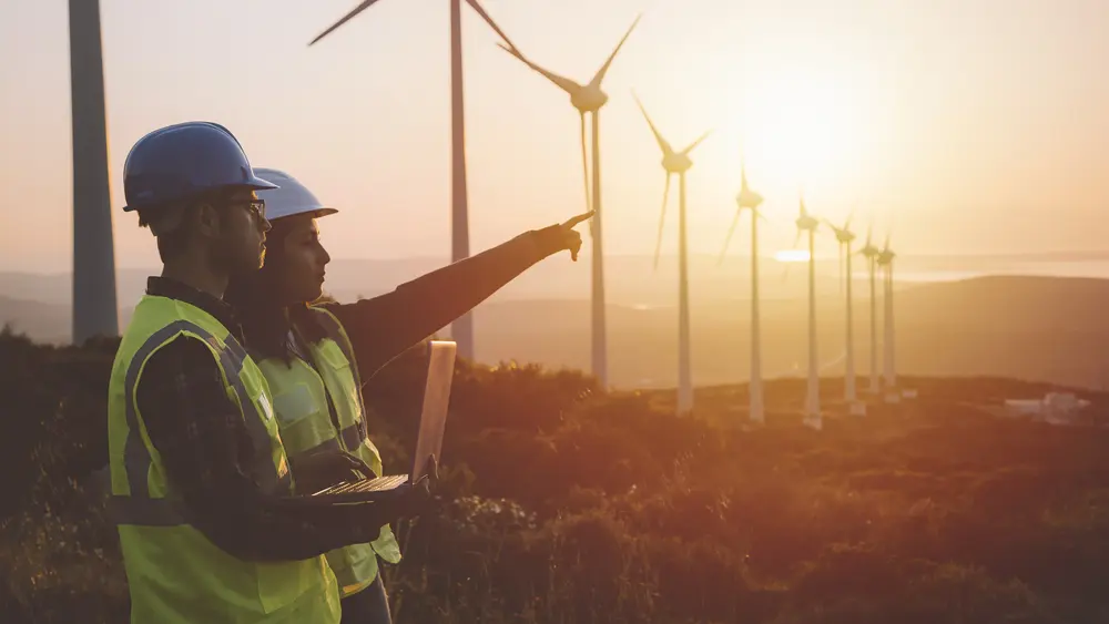 Zwei Personen mit Schutzhelmen zeigen auf Windkraftanlagen in hügeliger Landschaft bei Sonnenuntergang.