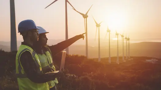 Bau und Betrieb regionaler Windparks Zwei Personen mit Schutzhelmen zeigen auf Windkraftanlagen in hügeliger Landschaft bei Sonnenuntergang.