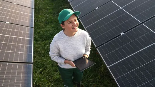 Photovoltaik für Unternehmen, Gewerbe und Landwirtschaft Eine Frau läuft durch ein Feld mit Photovoltaikanlagen mit einem Tablet in der Hand und lächelt.