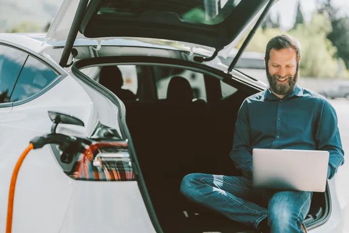 Flexibler Ladezugang für E-Fahrzeuge Eine Person sitzt im geöffneten Kofferraum eines weißen Elektroautos und arbeitet am Laptop, während das Auto lädt.