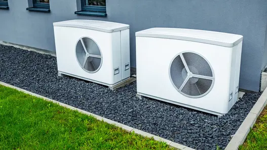 Effiziente Wärmepumpen für Wohngebäude Zwei weiße Wärmepumpen stehen auf einem Kiesbett neben einer Hauswand und sind betriebsbereit installiert.