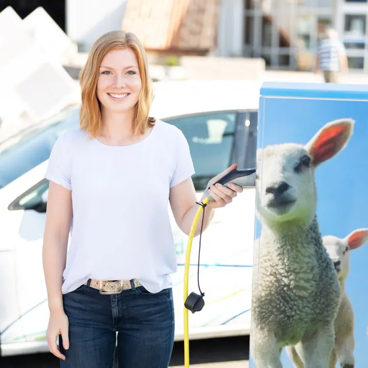 Kreative Ladesäulendesigns für Unternehmen Eine Person verbindet ein Elektroauto mit einer Ladesäule, die mit einem Schafmotiv und E-Auto-Symbol gestaltet ist.