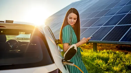 E-Mobilität in Kombination mit Photovoltaik Eine Person steht mit Smartphone neben einem Elektroauto, das bei Sonnenuntergang mit Solarstrom geladen wird.
