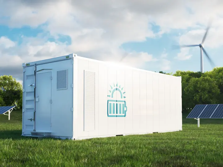 Innovative Batteriegroßspeicher Ein weißer Batteriegroßspeicher mit steht vor Solarpaneelen und einem Windrad auf einer Wiese.