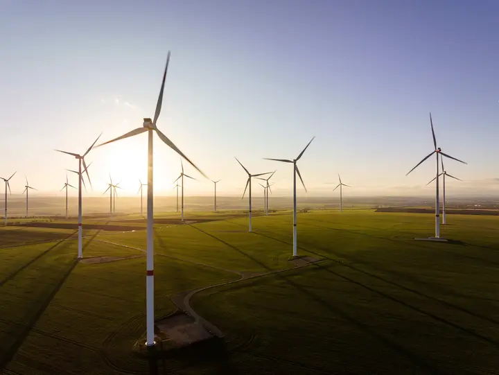 Batteriespeicher für die Integration erneuerbarer Energien Mehrere Windräder stehen auf einem grünen Feld, während die tiefstehende Sonne lange Schatten wirft.