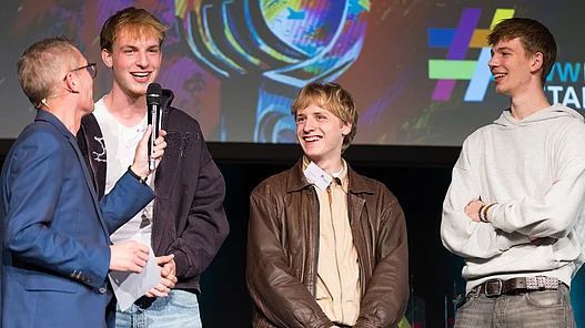 Band Motion - Start-Bonus Die Band Motion hält eine Urkunde für den Start-Bonus des WWKulturSTARTER26 in der Hand, während der Moderator Ingo Börchers ein Interview auf der Bühne führt.