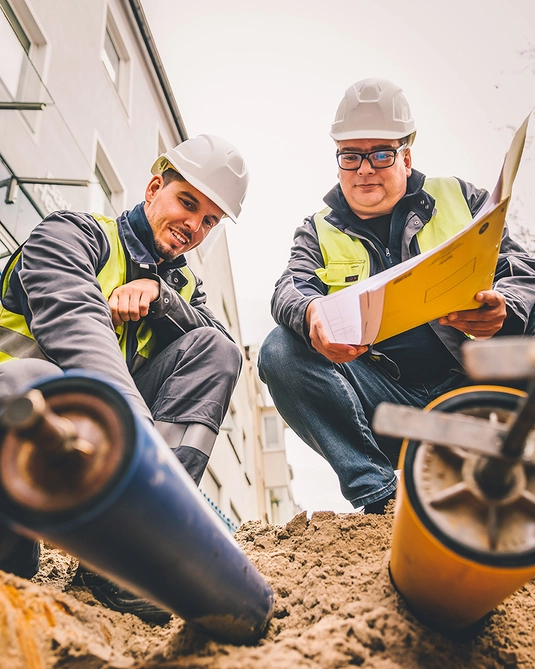 Bild mit zwei Personen kniend auf einer Baustelle in Arbeitskleidung mit Warnweste 