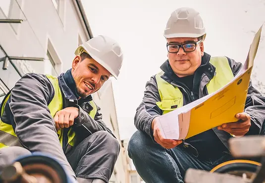 Bild mit zwei Personen kniend auf einer Baustelle in Arbeitskleidung mit Warnweste 