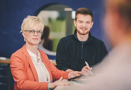 zwei Personen sitzend am Tisch mit Blick in die Kamera im Vordergrund eine optisch verschwommene Persoon