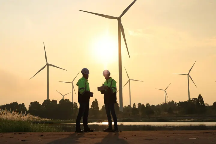 Netzausbau zum Gelingen der Energiewende Zwei Fachkräfte in Sicherheitskleidung arbeiten vor Windrädern in einer sonnigen Landschaft zusammen.