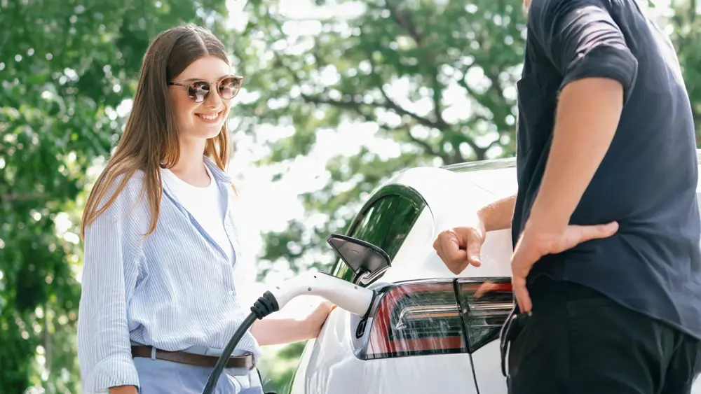 Zwei Personen stehen neben einem weißen Elektroauto, das im Freien über ein Ladekabel geladen wird.