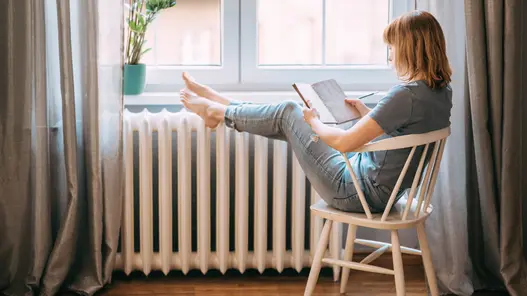 Lesepause Eine Frau sitzt auf einem Stuhl am Fenster, liest ein Buch und hat die Füße auf der Heizung abgelegt