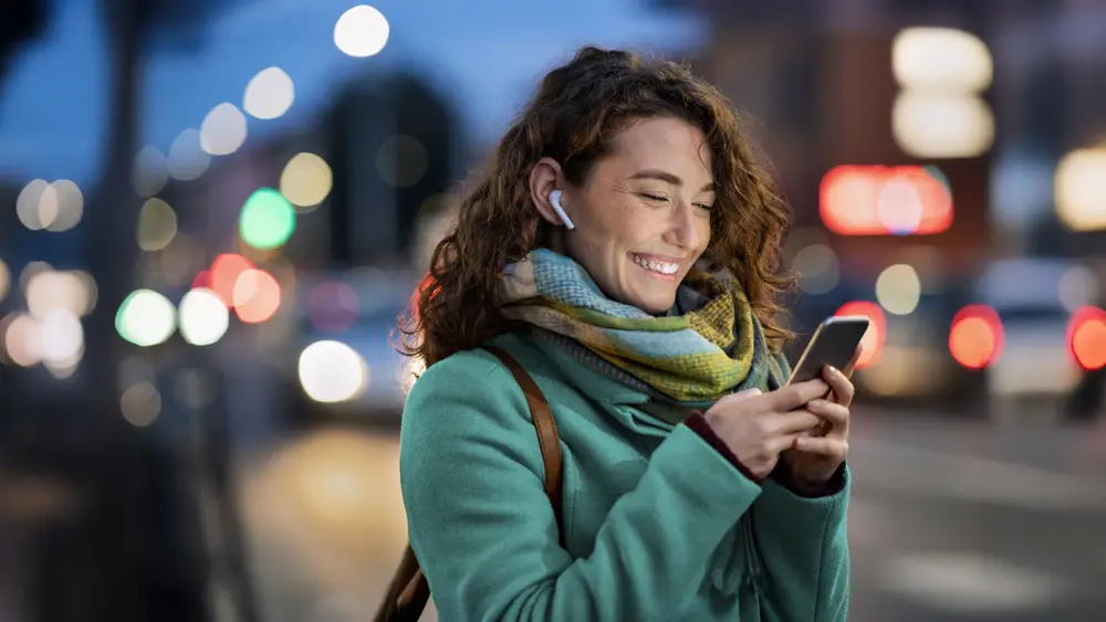 Eine Person mit Smartphone und Kopfhörern steht abends in einer beleuchteten Stadtumgebung mit verschwommenem Straßenhintergrund.